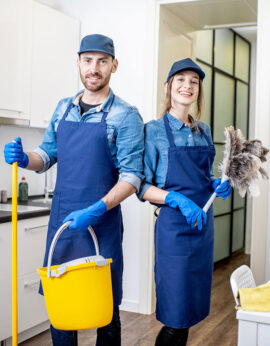 Portrait of a couple as a professional cleaners in uniform standing together with cleaning tools indoors